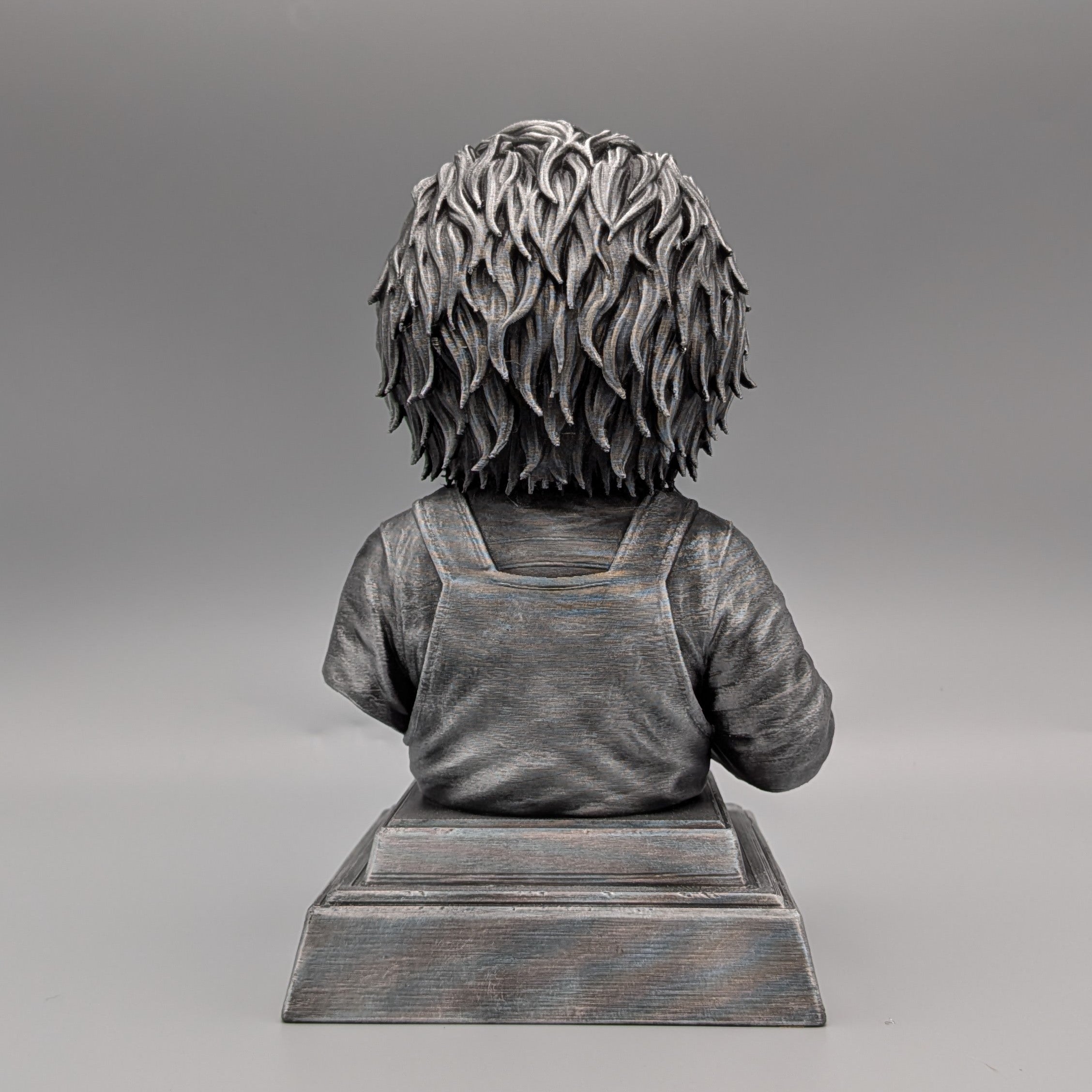 Rear view of a silver Chucky-inspired horror doll bust, showing sculpted hair, textured overalls and a pedestal base.