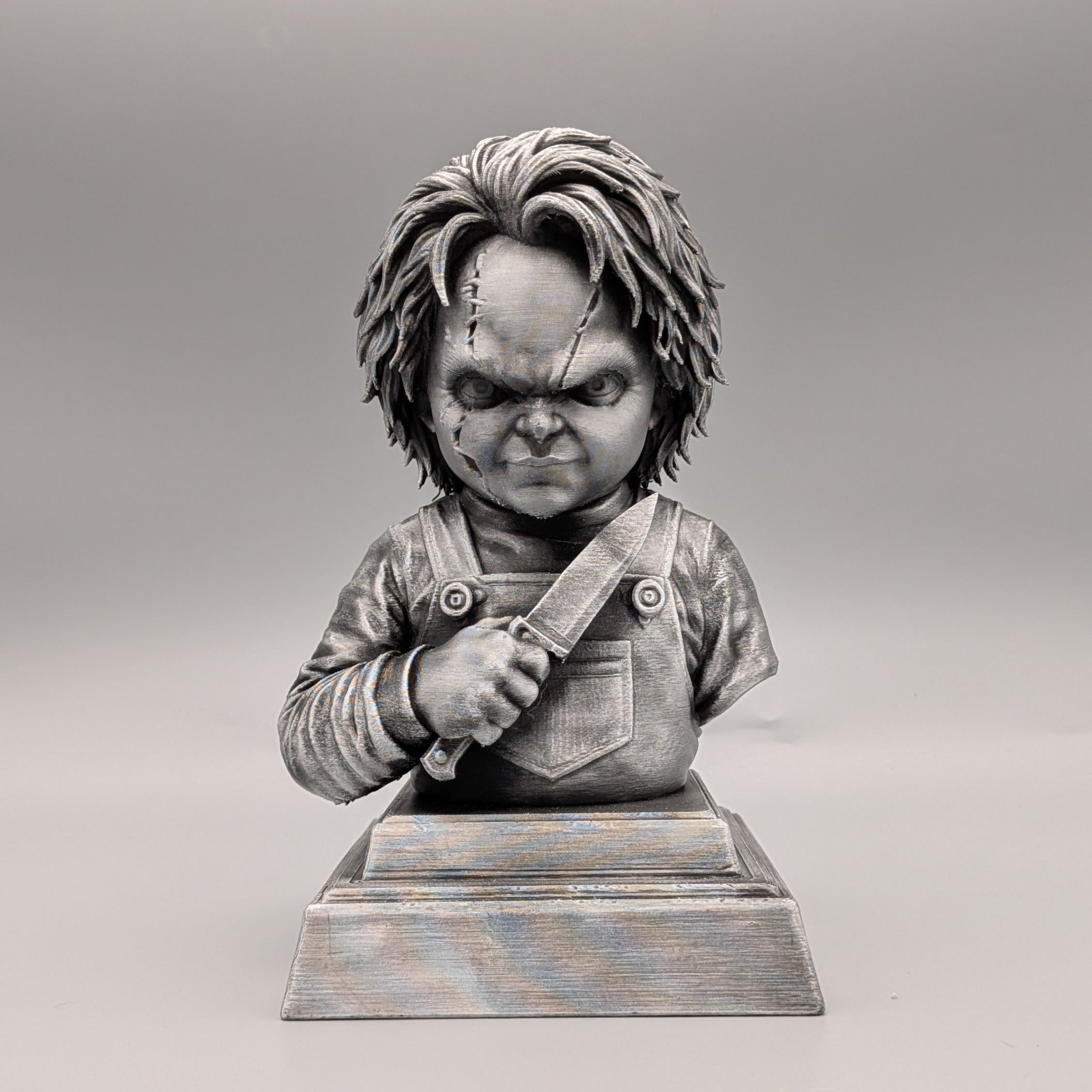 Front view of a silver Chucky-inspired horror doll bust holding a knife, with facial scars, overalls and a detailed pedestal base.