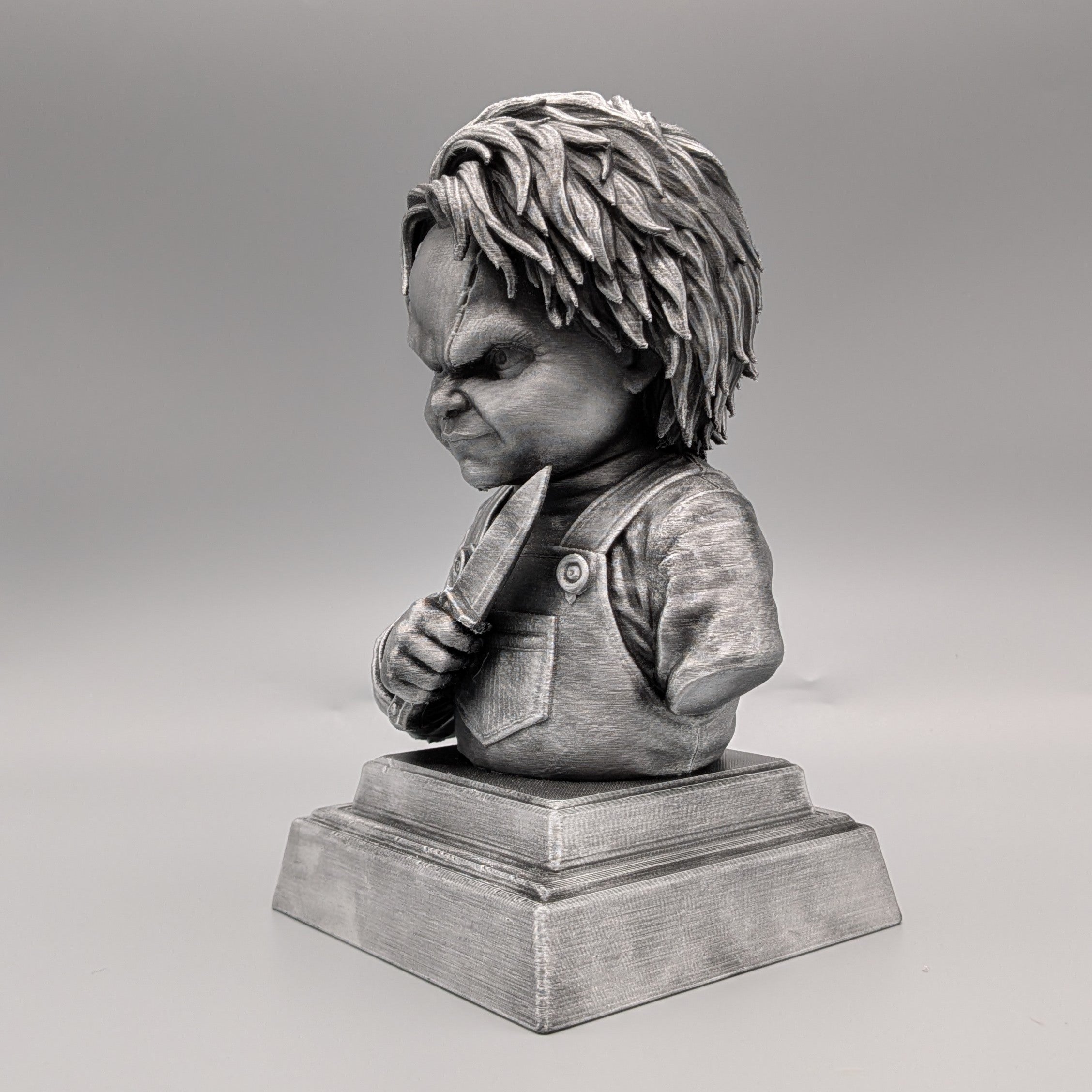 Left-side view of a silver Chucky-inspired horror doll bust holding a knife, featuring sculpted hair, overalls and facial scars.