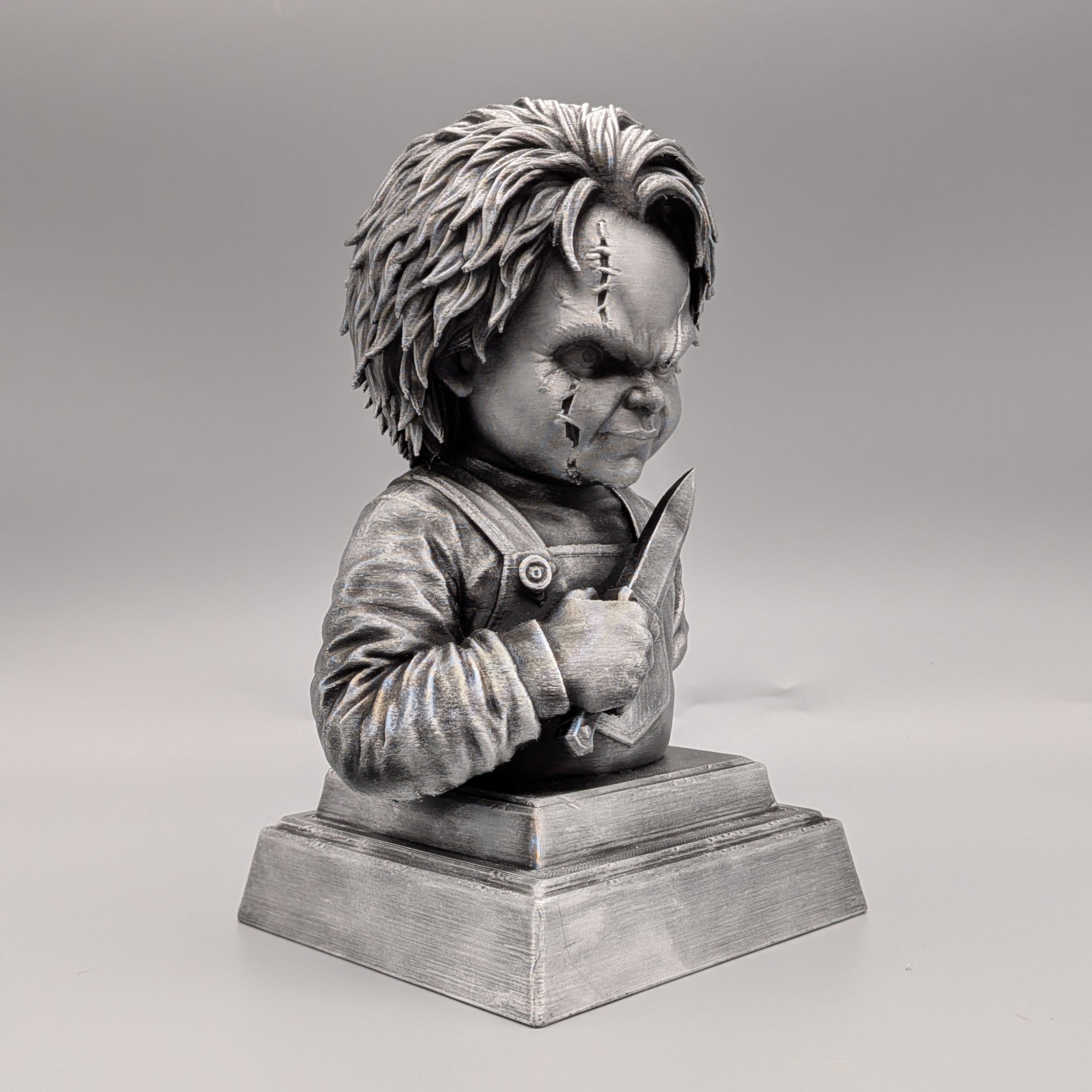 Right-side view of a silver Chucky-inspired horror doll bust holding a knife, with detailed facial scars, clothing and pedestal base.