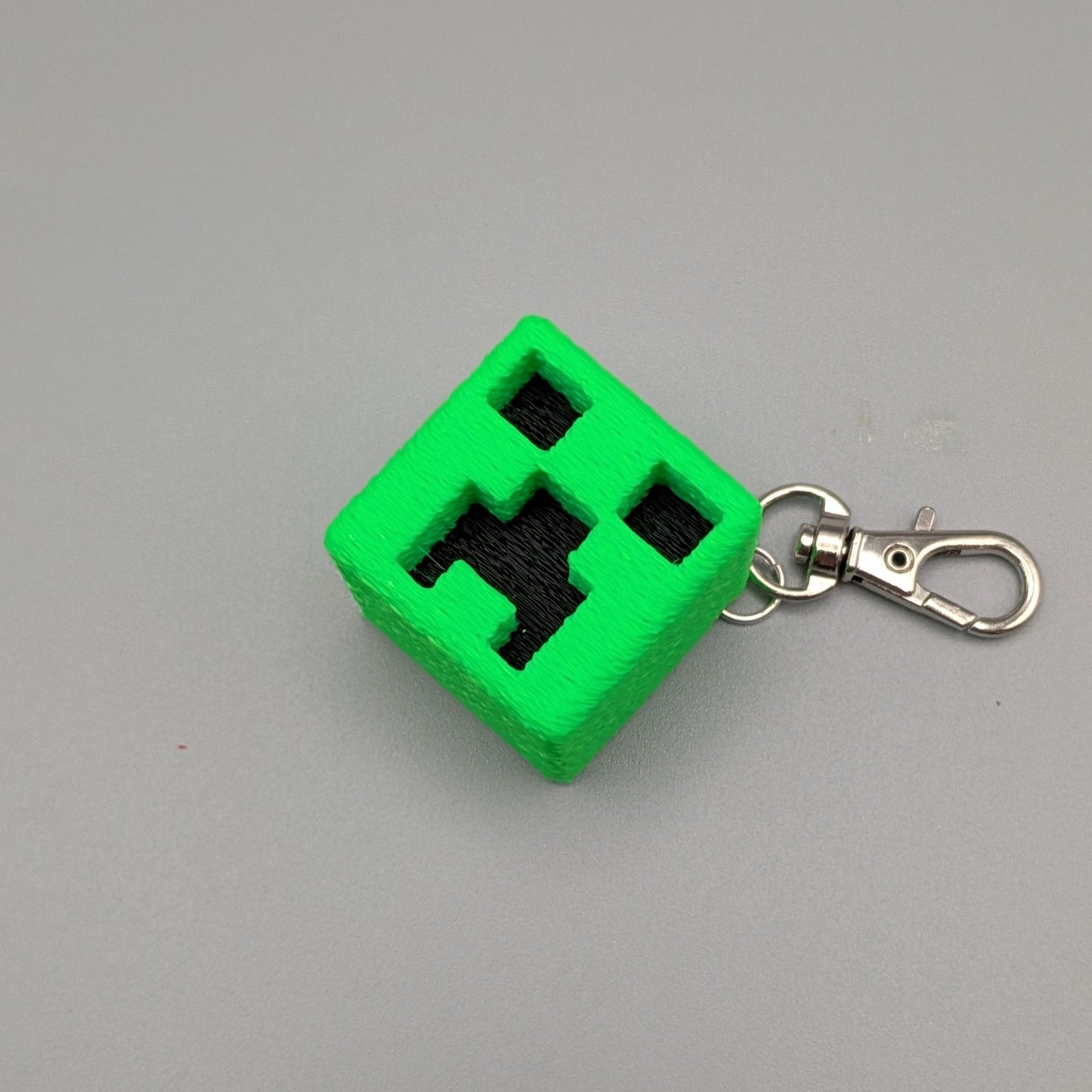 Angled top view of a green Creeper-inspired cube keychain showing the black pixel face and metal clasp