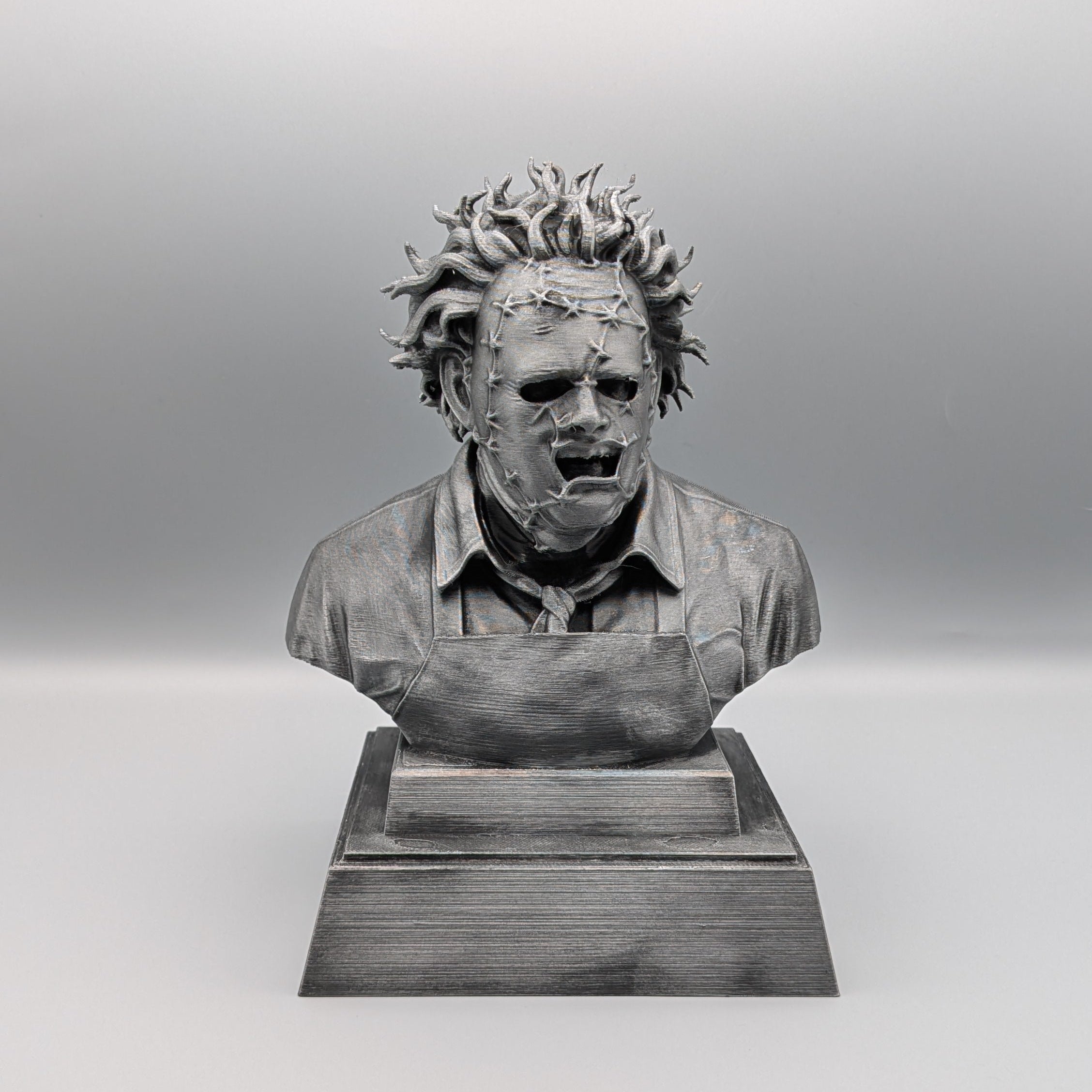 Front view of a 20 cm hand-painted silver Leatherface tribute bust with a stitched mask and apron on a pedestal base