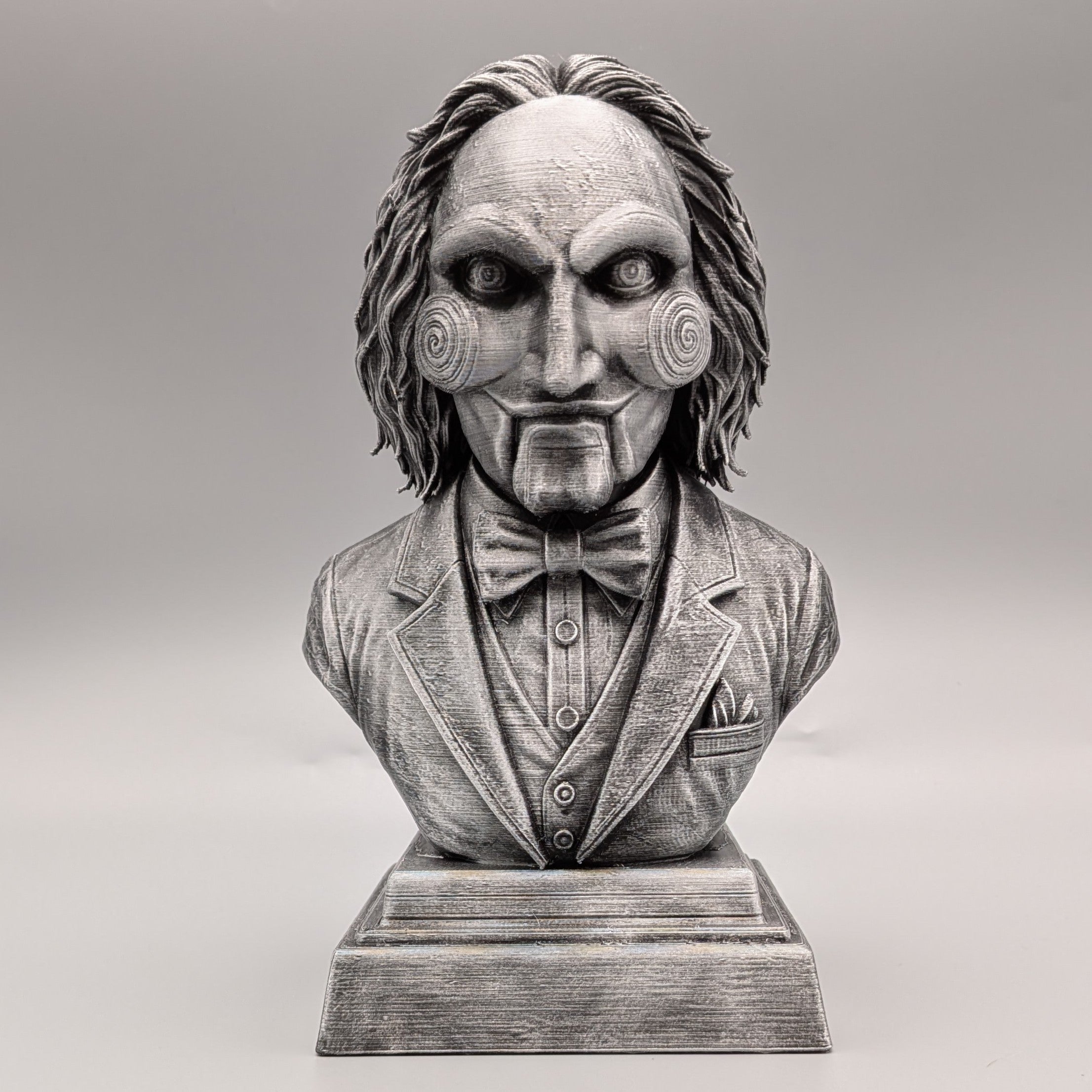 Front view of a silver Saw-inspired horror puppet bust with spiral cheeks, suit, bow tie and detailed pedestal base.