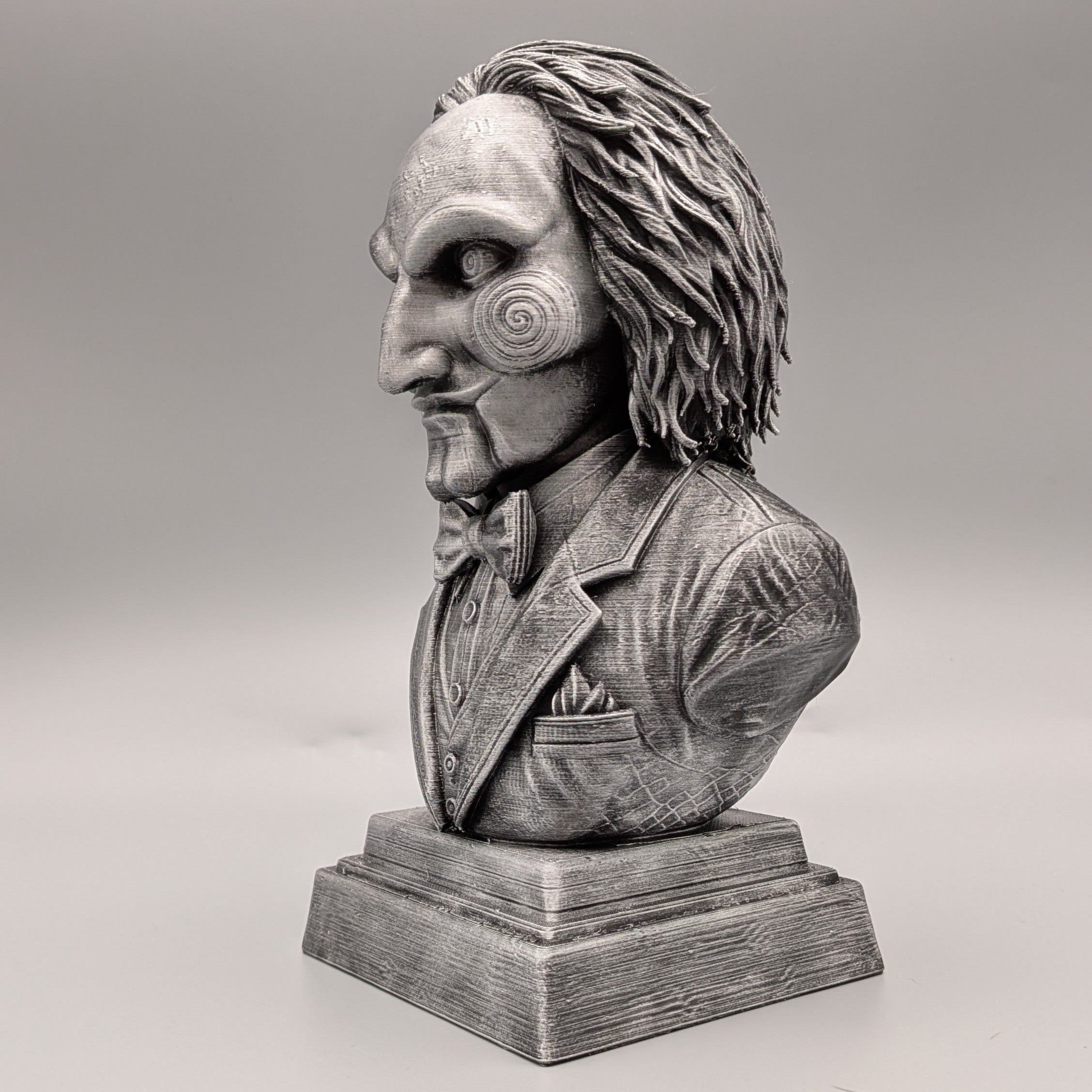 Left-side view of a silver Saw-inspired horror puppet bust with spiral cheek, sculpted hair and detailed formal suit.