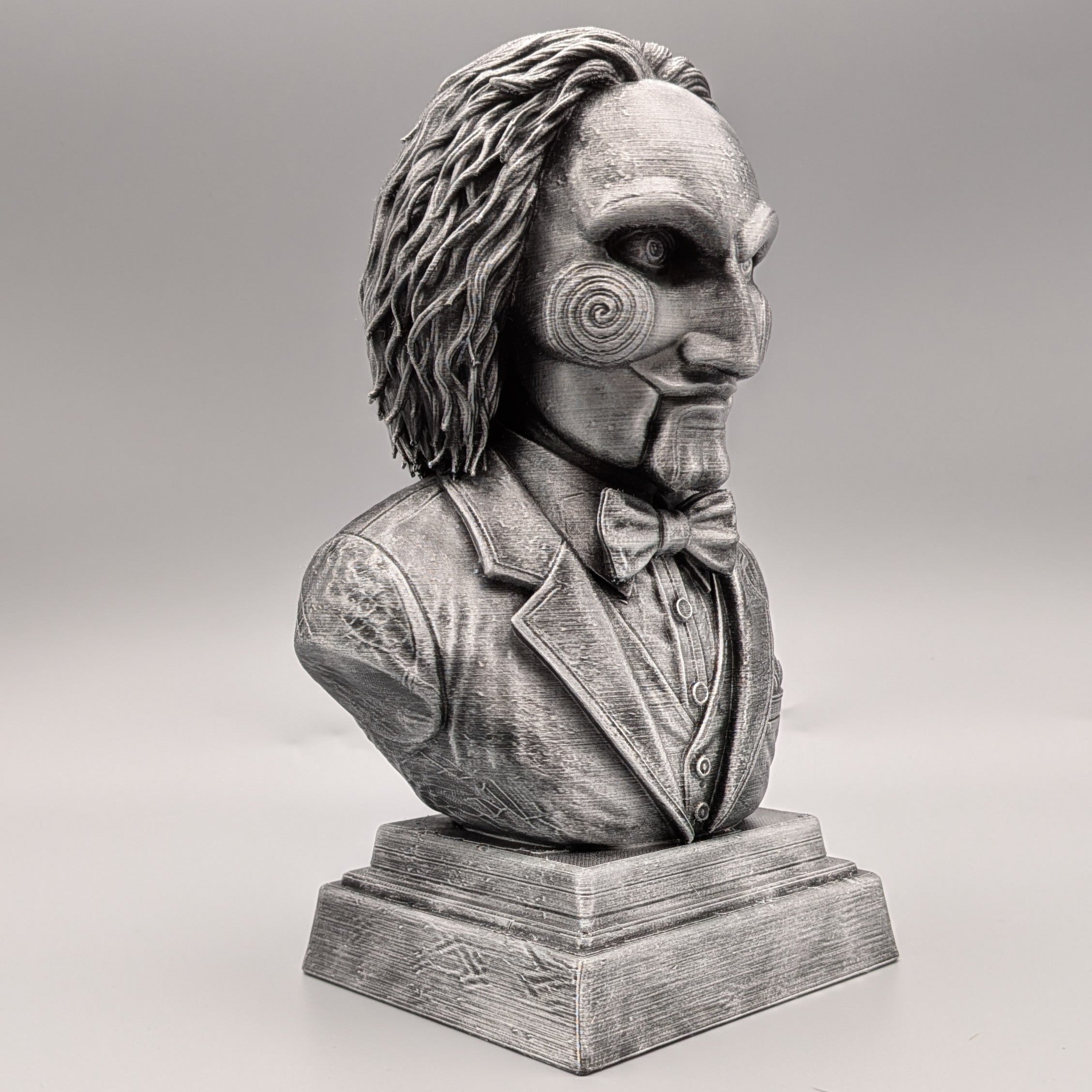 Right-side view of a silver Saw-inspired horror puppet bust with spiral cheek, detailed suit, bow tie and pedestal base.