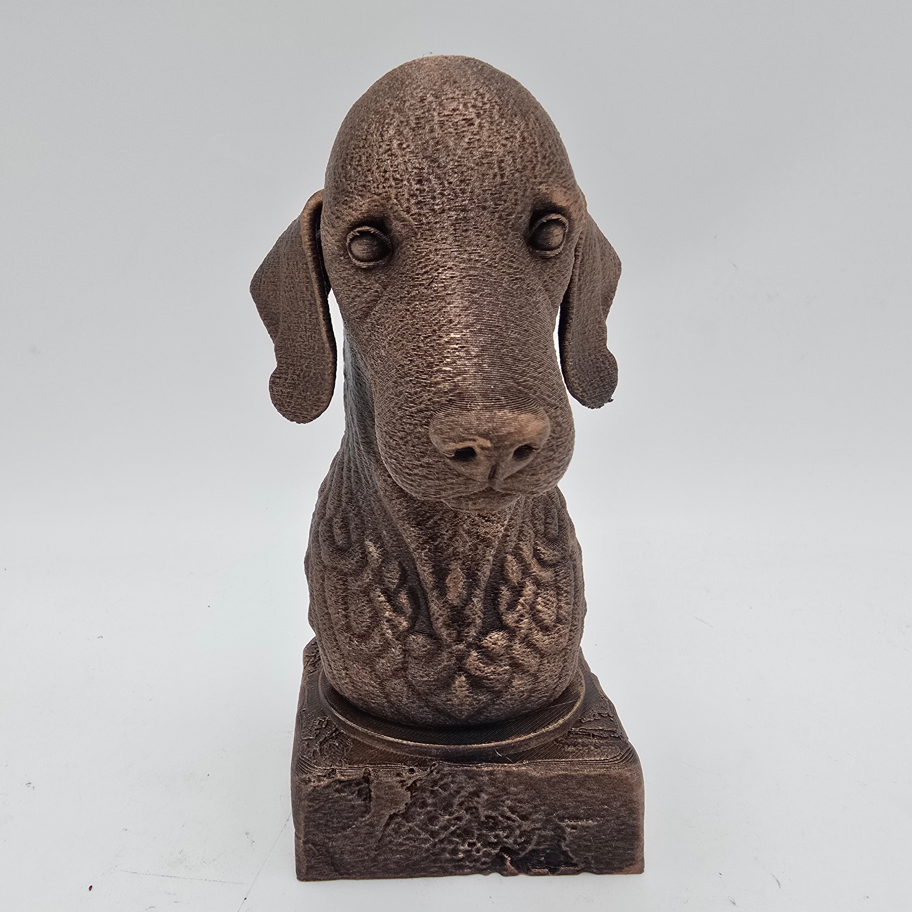 Vooraanzicht van een bronskleurige Bedlington terriër hondenbuste op een decoratieve sokkel tegen een witte achtergrond.