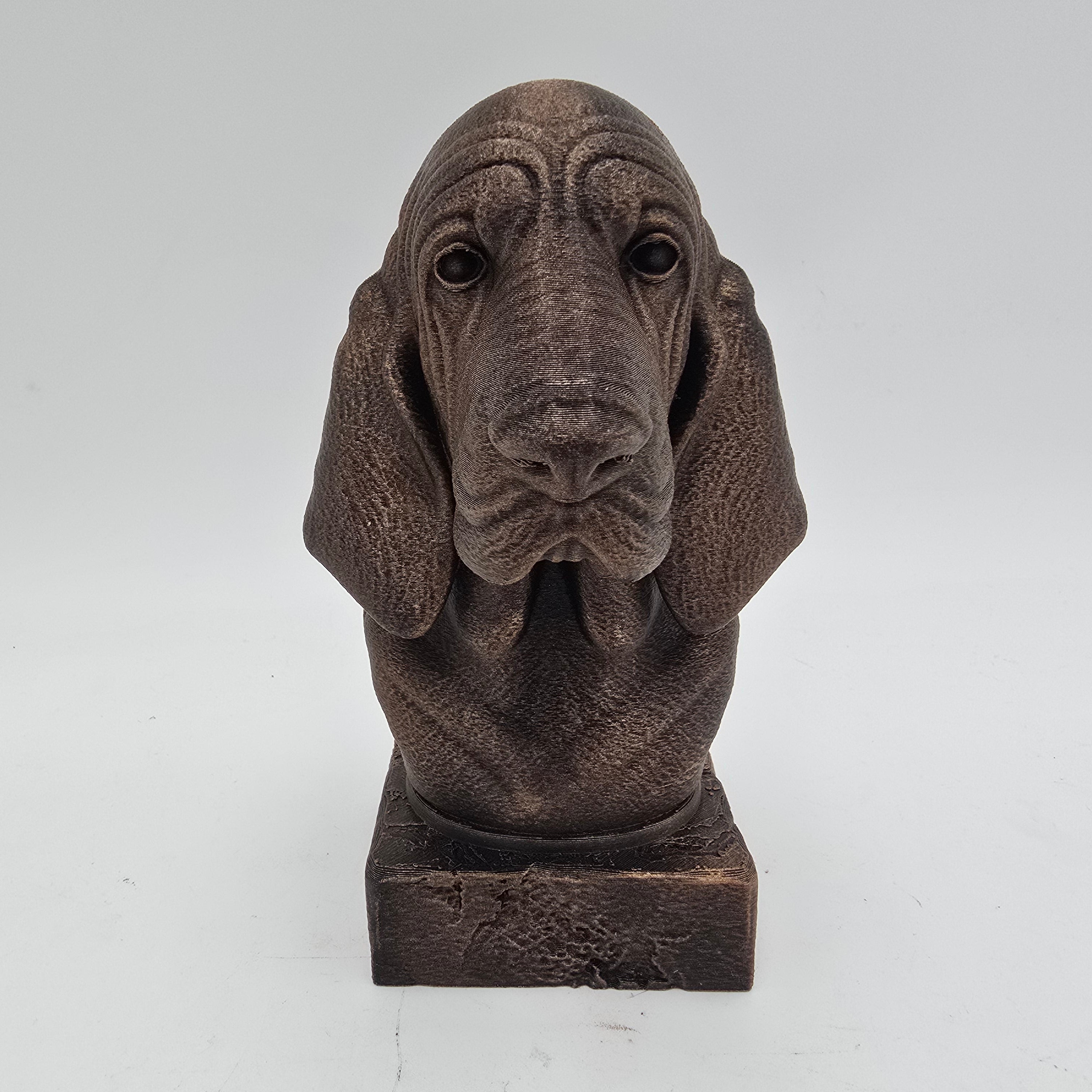 Front view of a bronze Bloodhound dog bust sculpture with long drooping ears and detailed facial folds on a decorative pedestal.